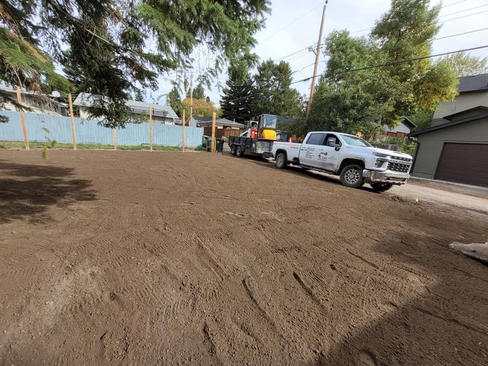 A white pickup truck with a trailer and a yellow skid-steer loader sits on a newly graded dirt lot by a residential home.