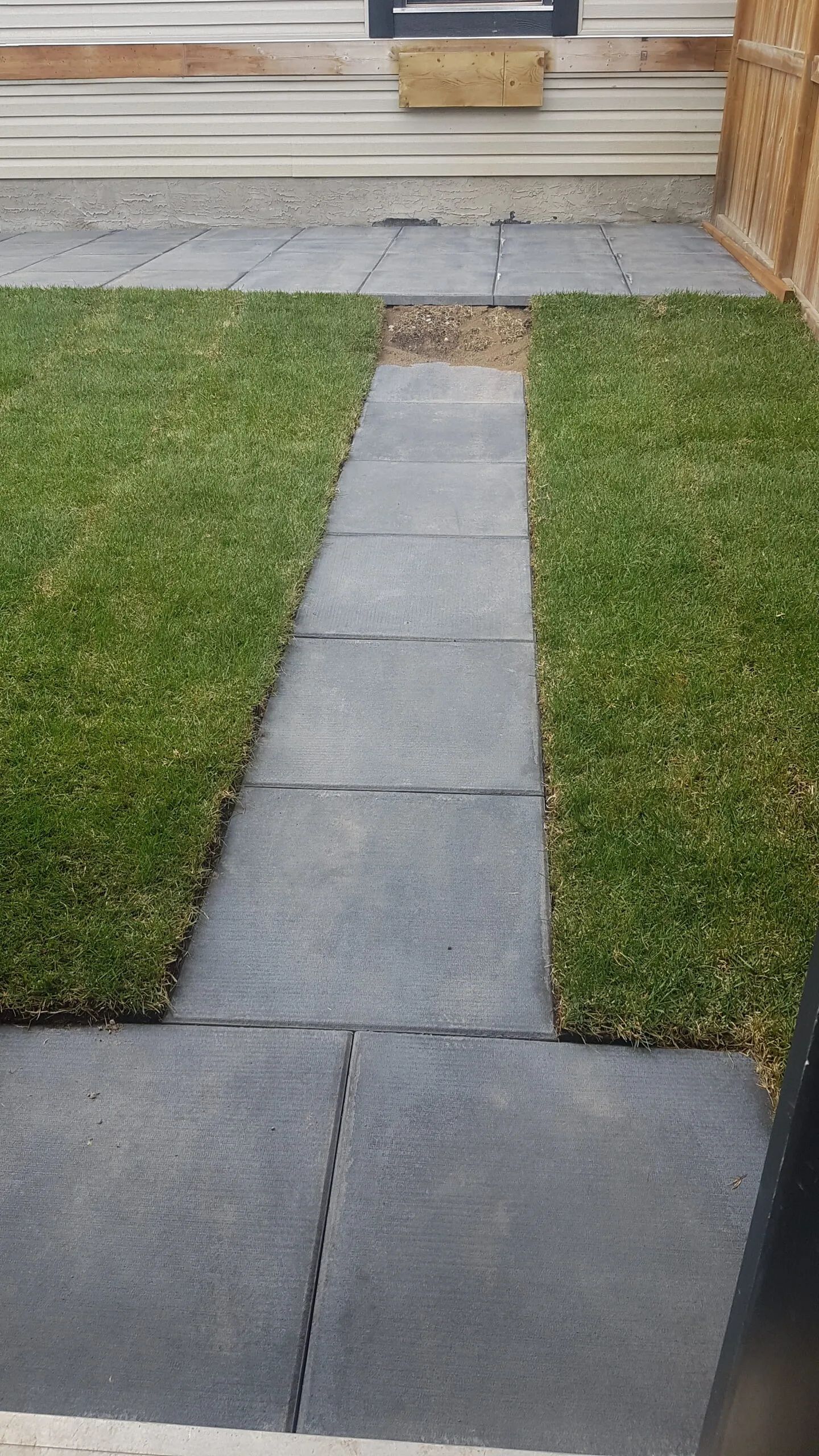 A stone paver path leads through a green lawn to a house exterior with light-colored siding and a wooden planter.