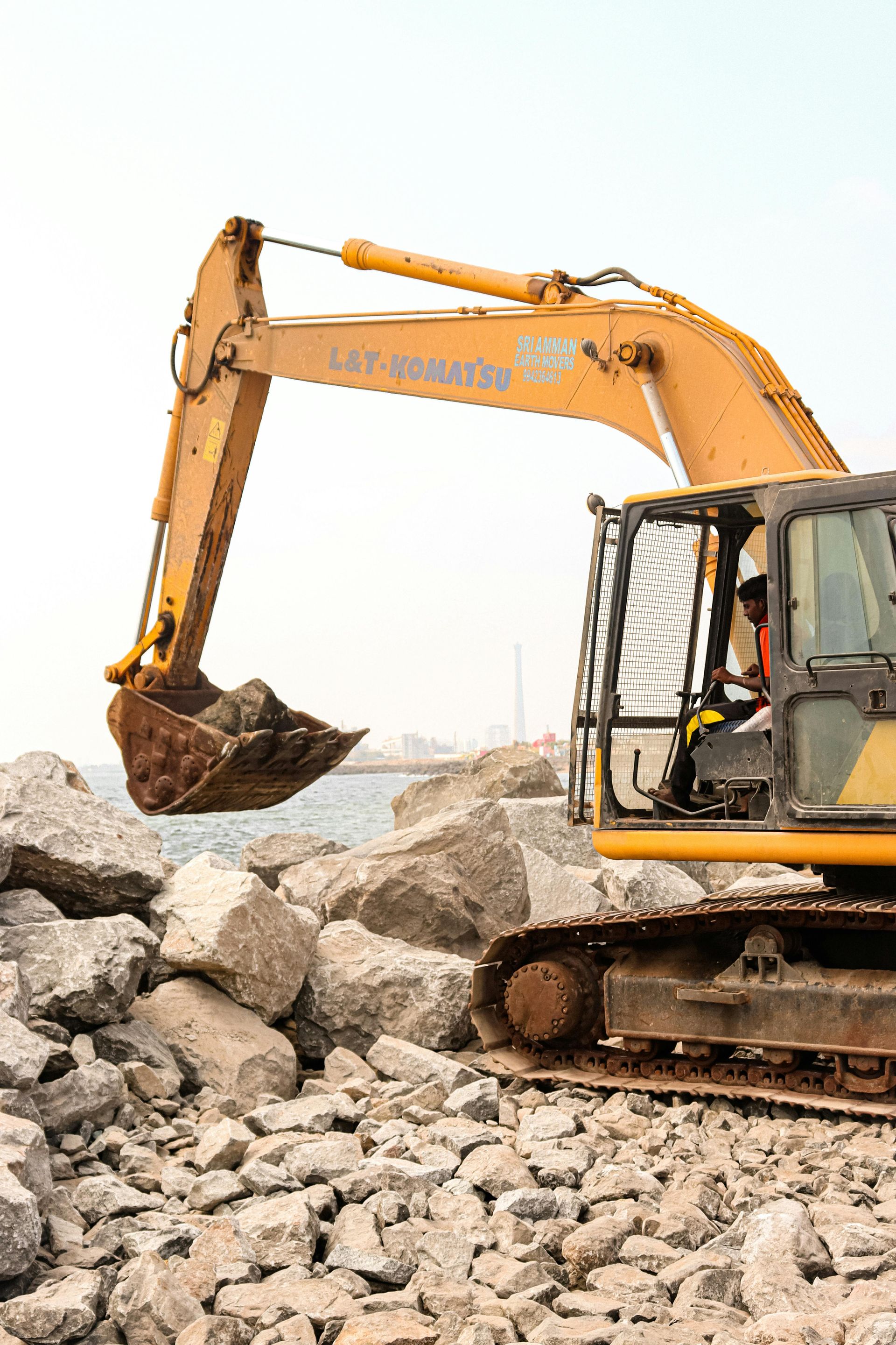 A yellow excavator lifts a large rock on a rocky shoreline near the water.