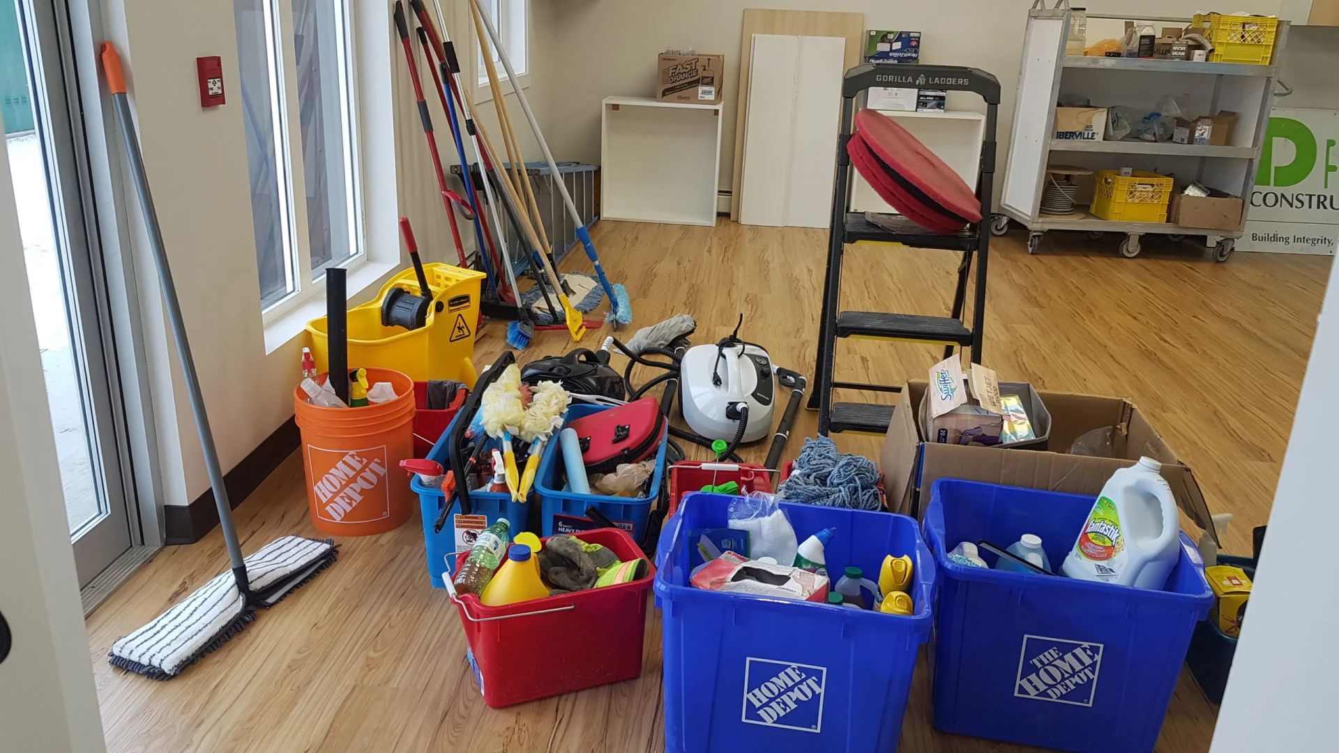A room containing cleaning supplies, including buckets, brooms, a step ladder, and containers of cleaning agents.