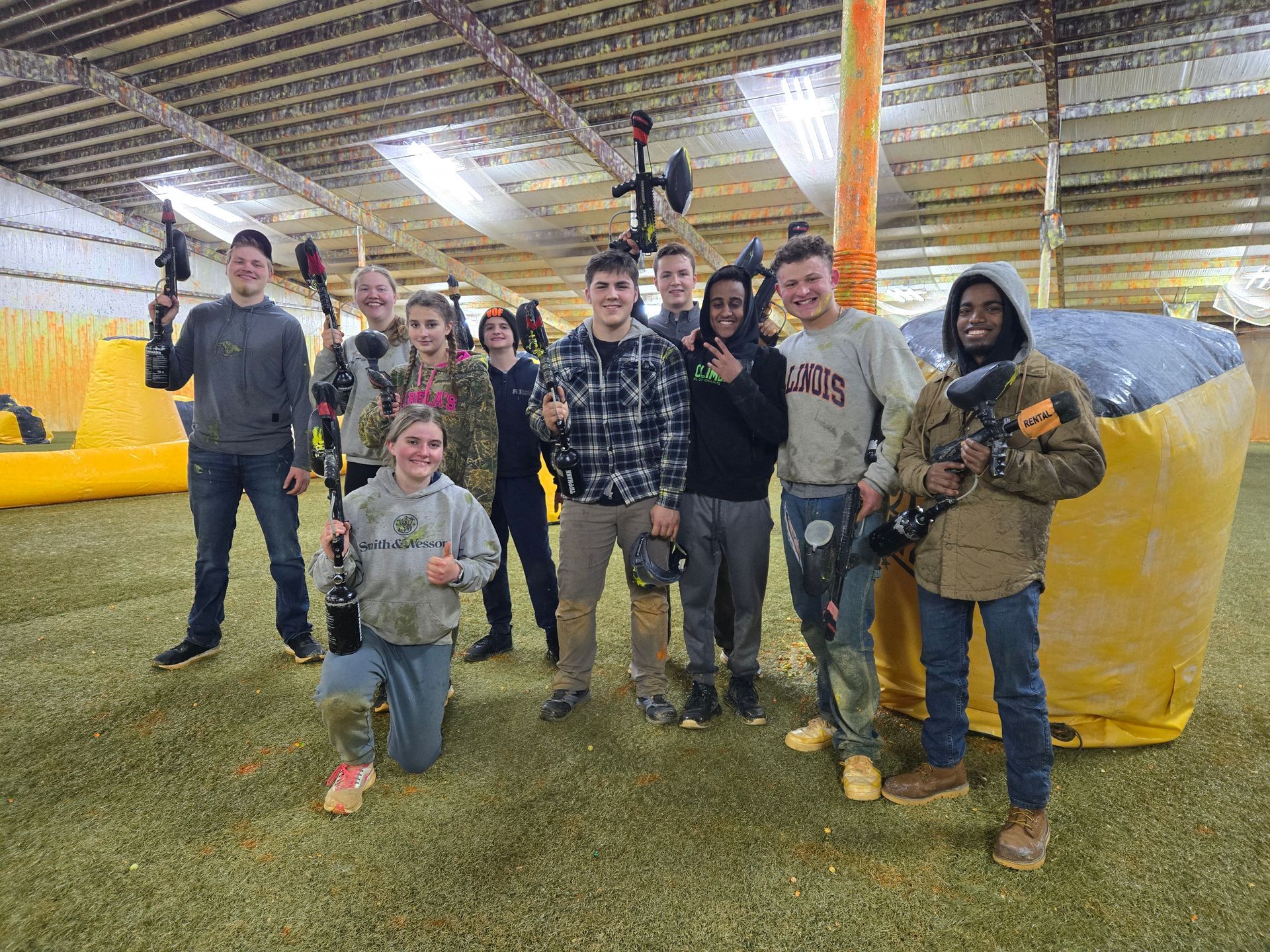 A group of people holding paintball guns posing on a green field inside a building. Most smile at the camera.