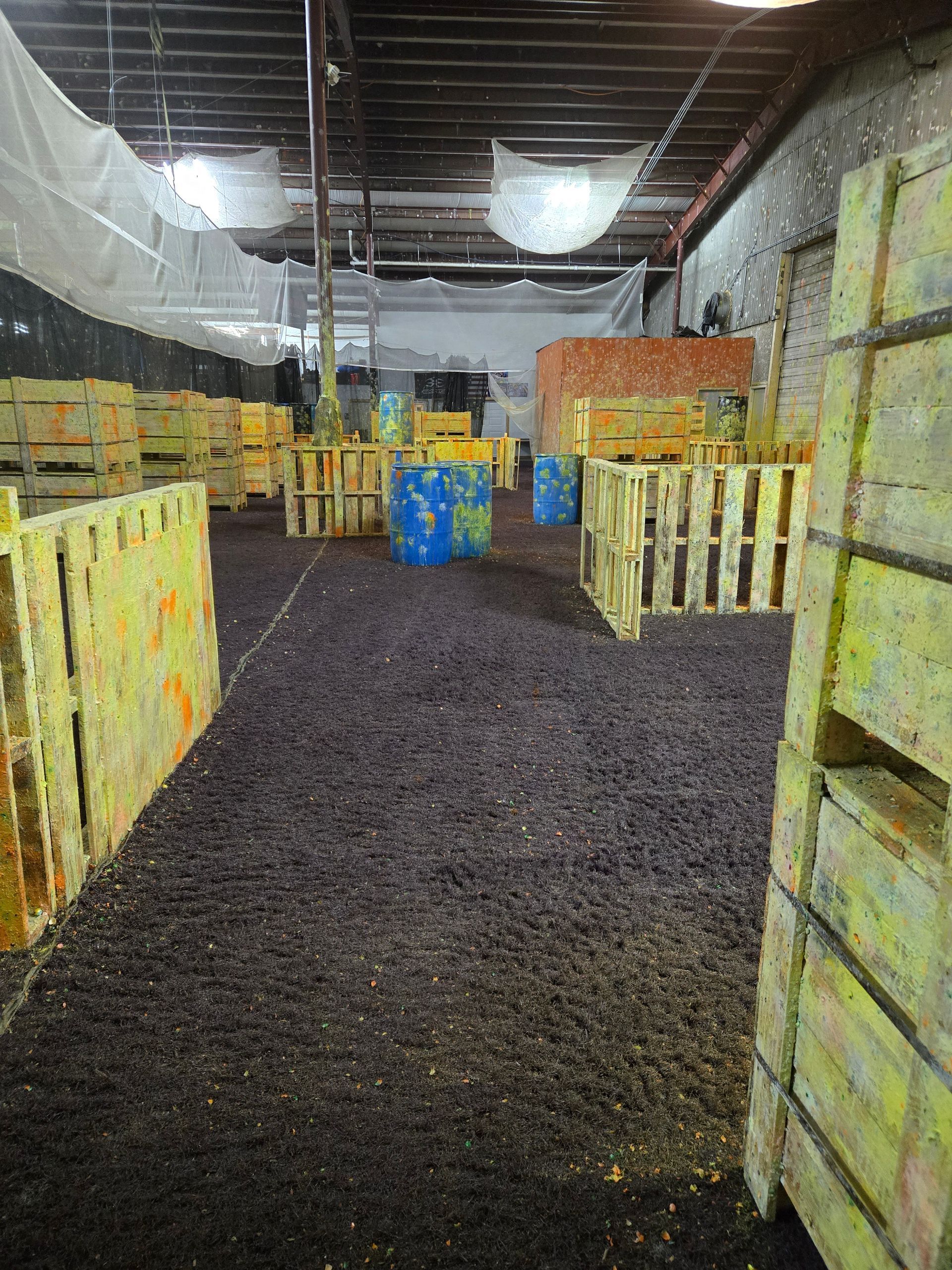 Indoor paintball arena with wooden barricades and a dark playing surface. Paint splatters cover the barriers.