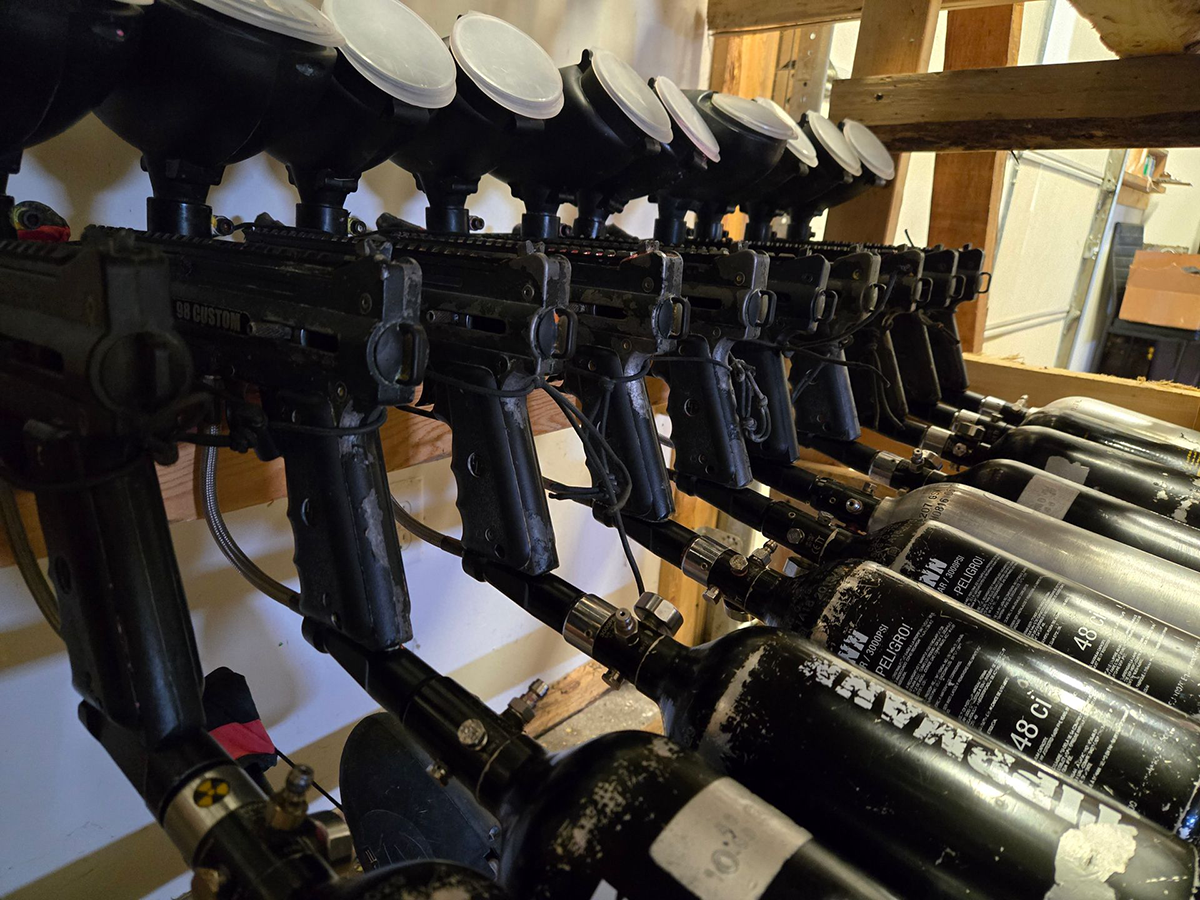 Row of black paintball guns with hoppers and air tanks, lined up on a rack.