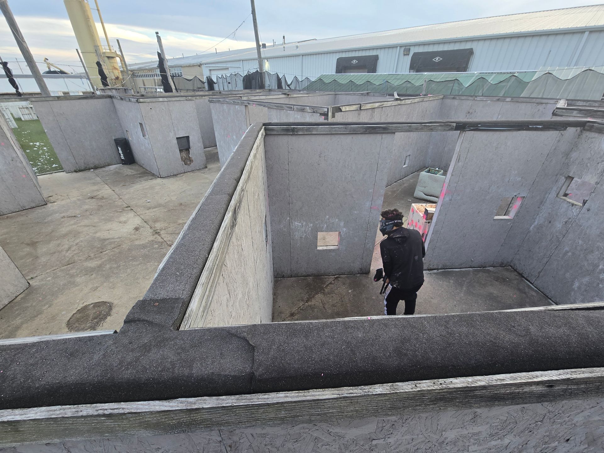 A person in black tactical gear aims a paintball gun from a concrete structure.