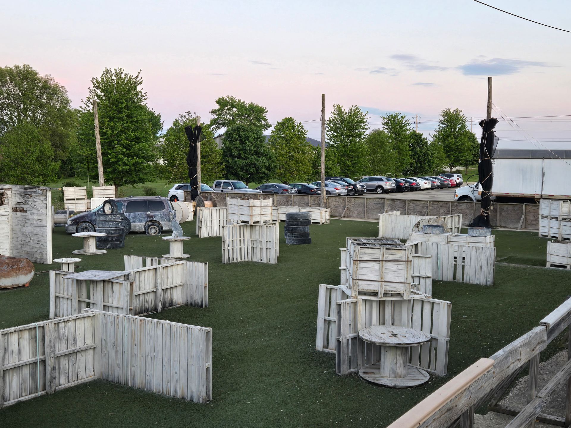Paintball field with white wooden barricades on green turf, cars in a distant parking lot, and a dusk sky.