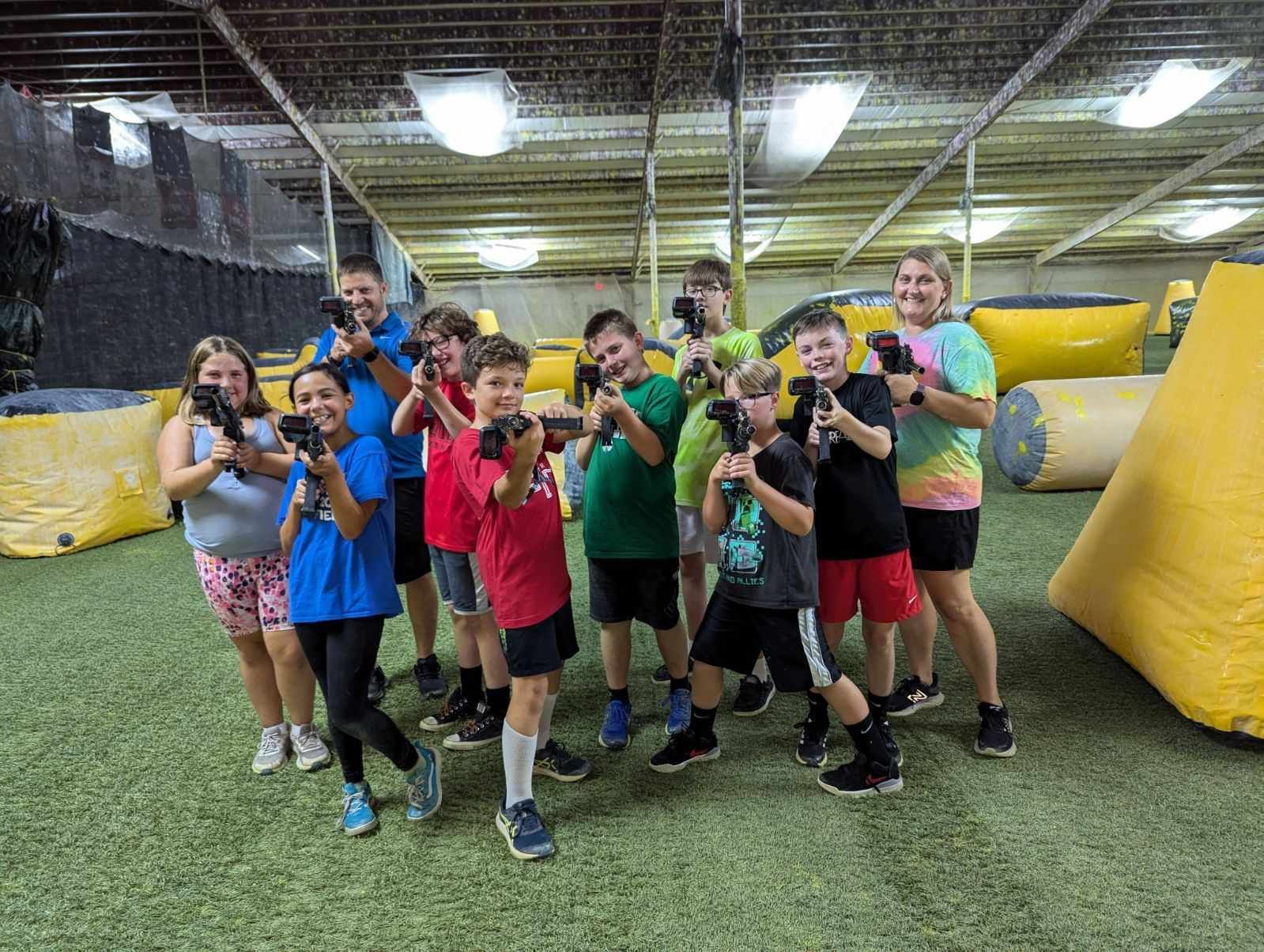 A group of kids and adults pose with laser tag guns in an indoor arena with inflatable barriers.
