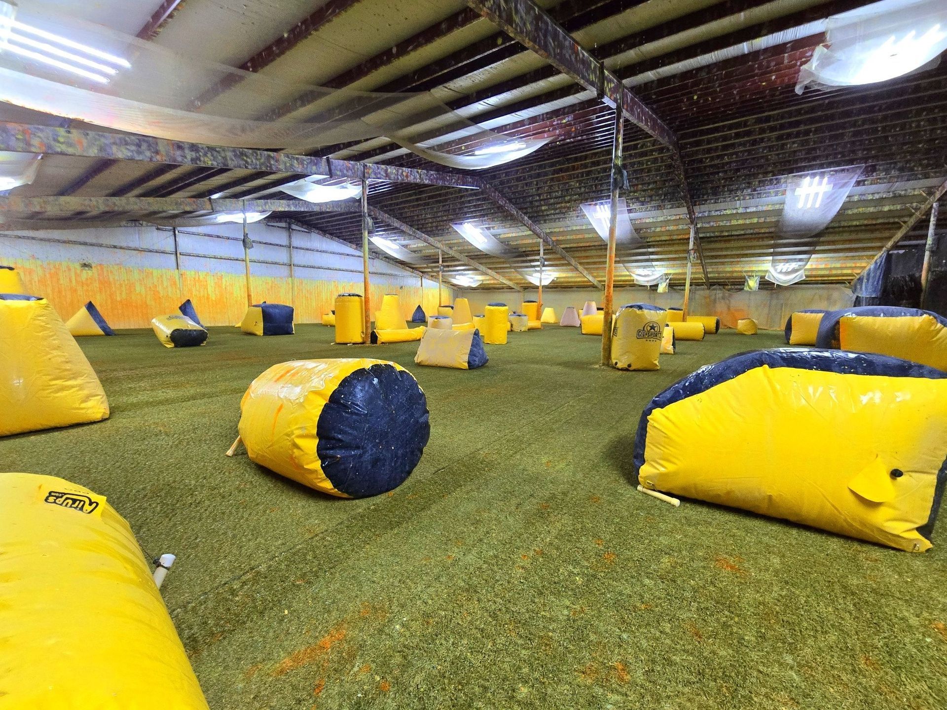 Indoor paintball arena with yellow and black bunkers on green turf. Fluorescent lights illuminate the space.