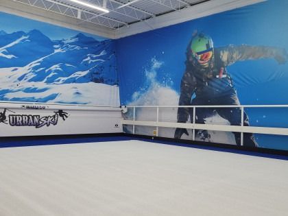 Indoor ski slope with blue and white backdrop. A skier is pictured on the wall and the floor is a simulated snowy surface.