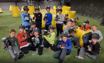 A group of children wearing helmets and holding paintball guns, posed in front of yellow bunkers on a paintball field.