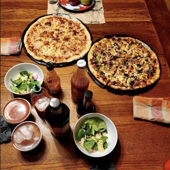 A wooden table is set for a meal with two pizzas, two salads, drinks, and various condiments.