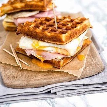 Waffle sandwich with egg, ham, and cheese on a wooden board, drizzled with sauce, held together with toothpicks.