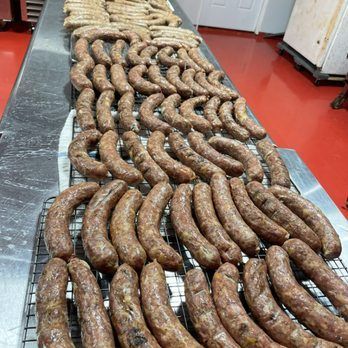 Rows of browned sausages cooling on wire racks, likely in a commercial kitchen. Red floor and stainless steel counter visible.