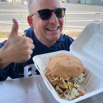Man in sunglasses smiles, giving a thumbs up while holding an open container with a large sandwich, outdoors.