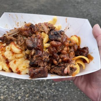 A hand holds a tray of barbecue: mac and cheese, pulled pork, baked beans, and brisket.