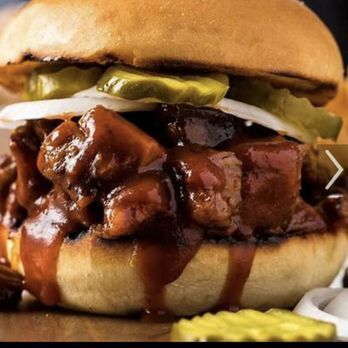 Close-up of a BBQ sandwich on a toasted bun, overflowing with saucy meat, pickles, and onions.