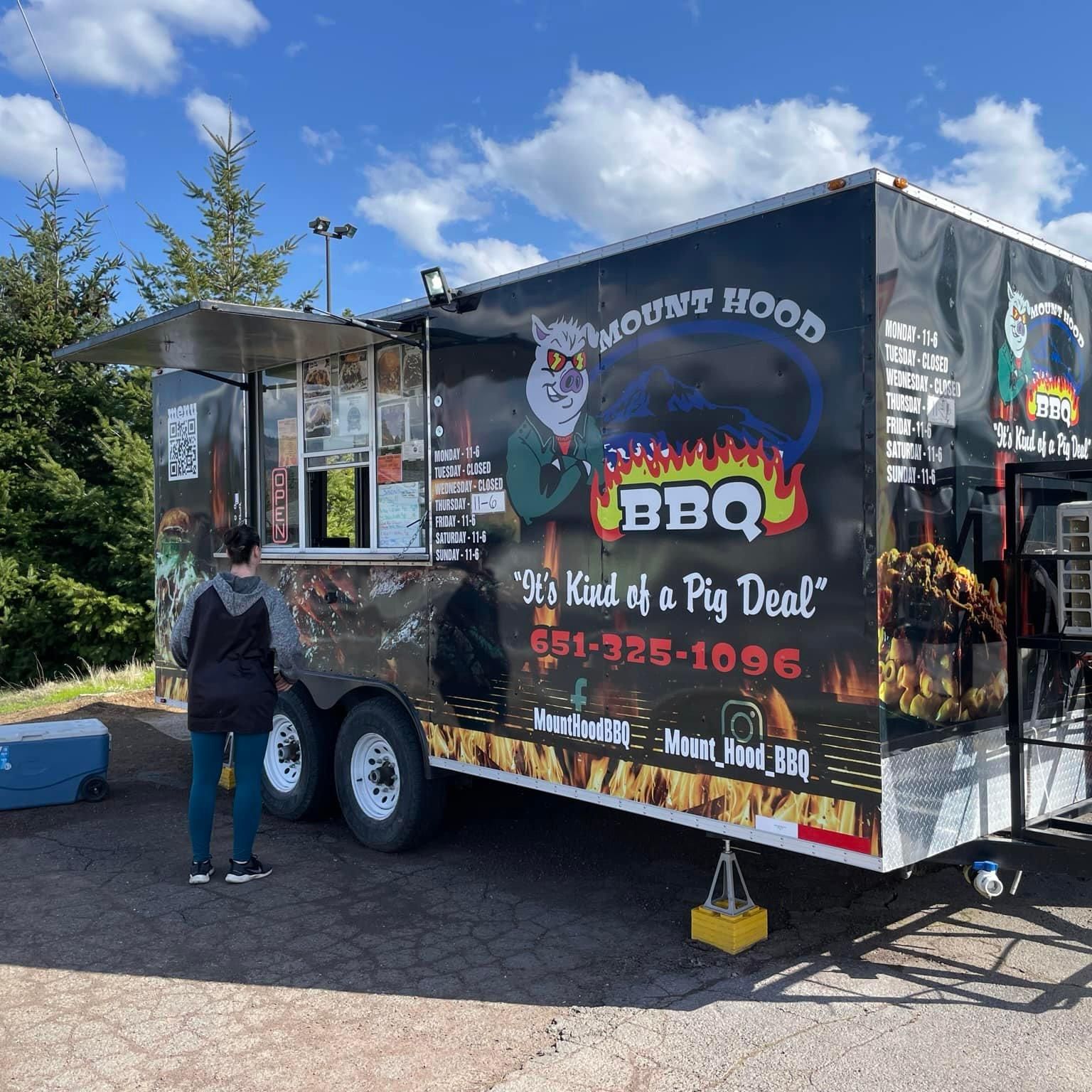 A person orders food from a Mount Hood BBQ food truck. The black truck features a logo of a pig wearing a hat, with flames and the tagline 