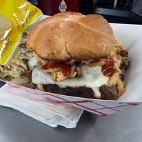 Bearded man in a suit excitedly bites into a burger, next to a bowl of fries.