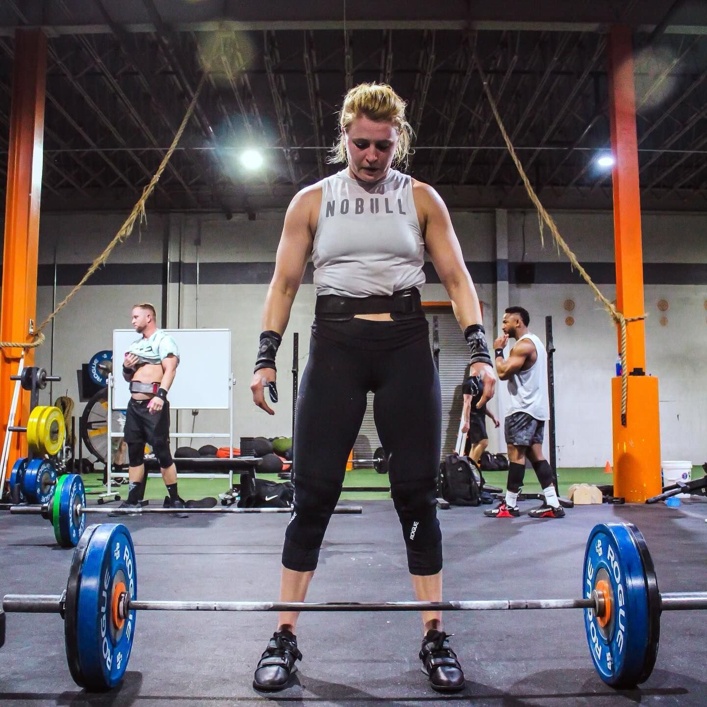 A woman is lifting a barbell in a gym wearing a nobull tank top