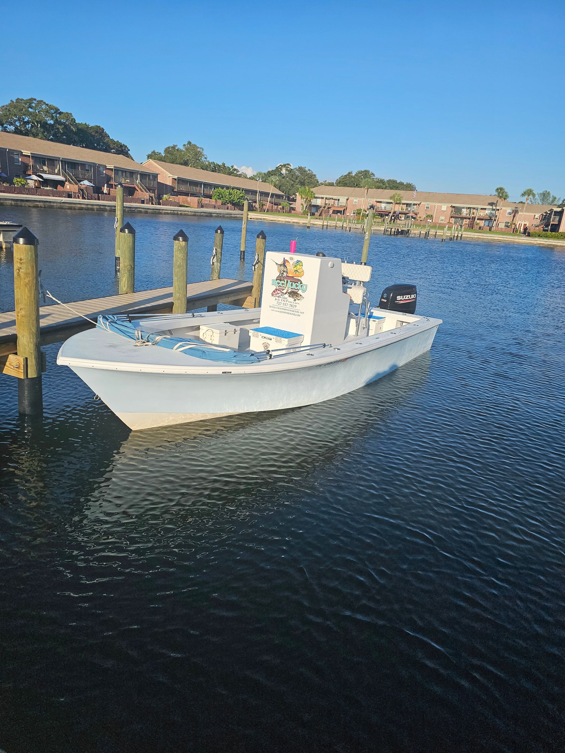 A white motorboat docked next to a wooden pier on a blue-water body. Buildings and clear sky in background.