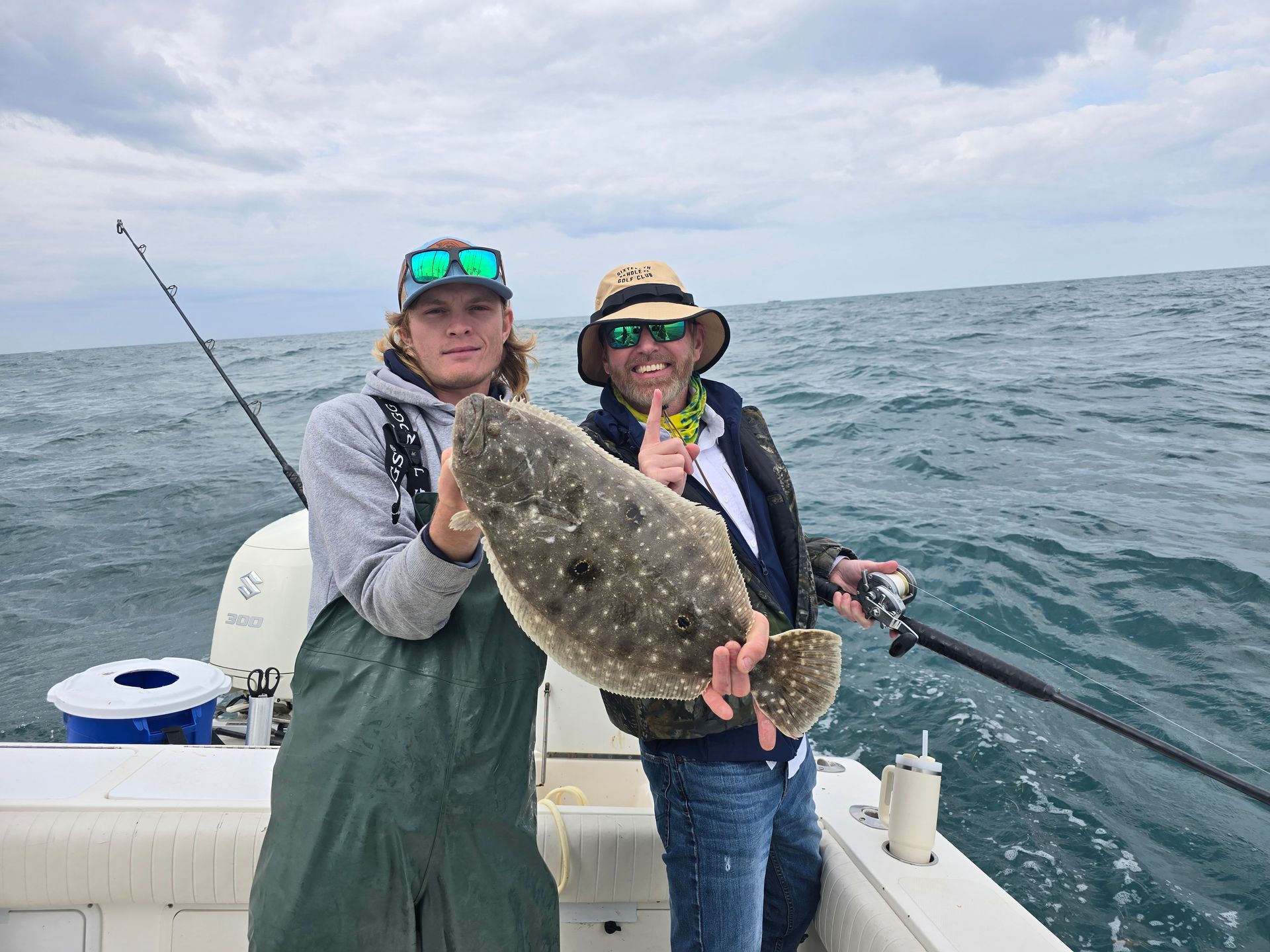 Two men on a boat holding a large, spotted flounder. They smile, the sky is overcast, and they are at sea.