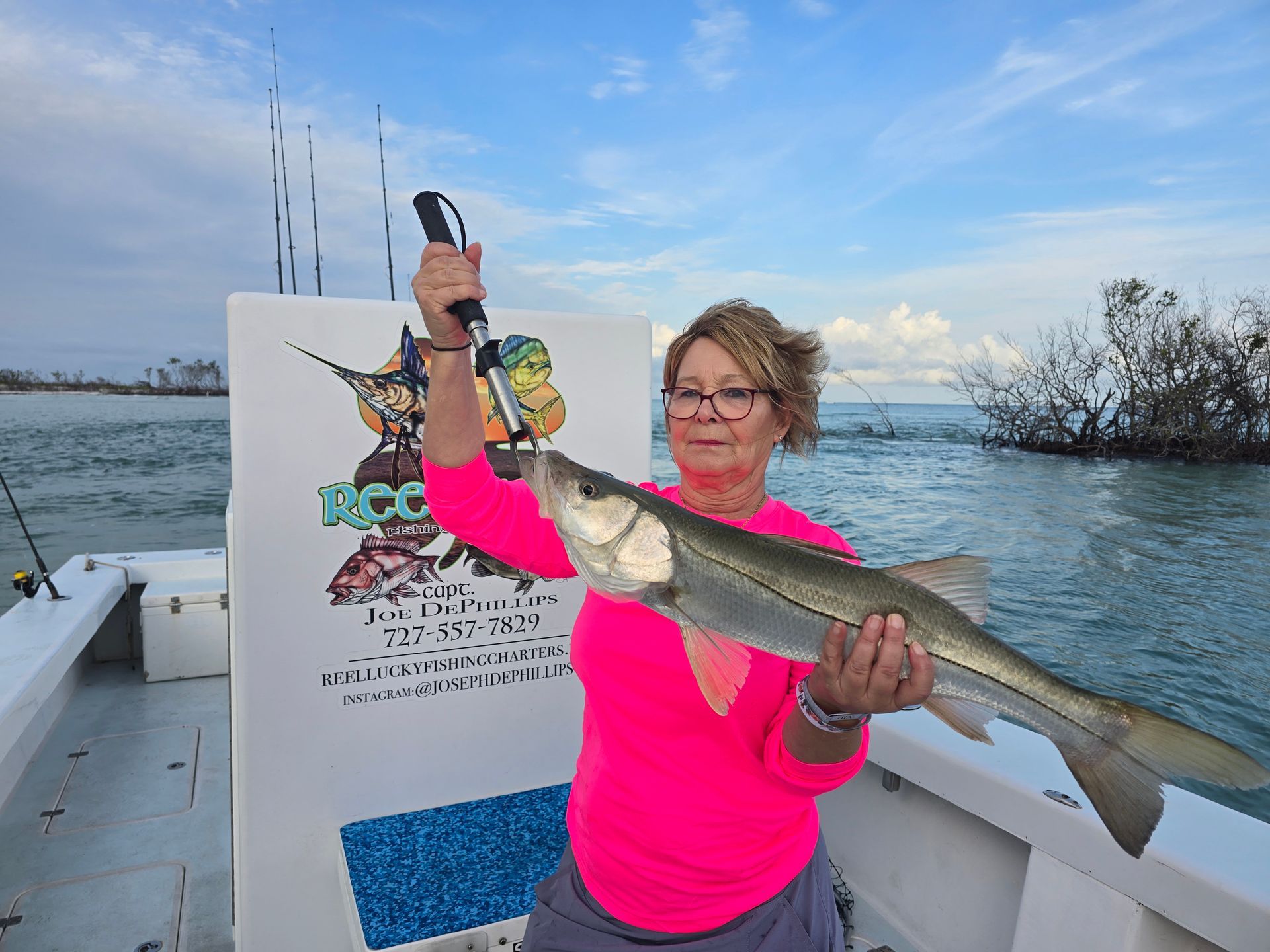 Woman holding a fish on a boat, arms raised, wearing pink, with a backdrop of water and sky.