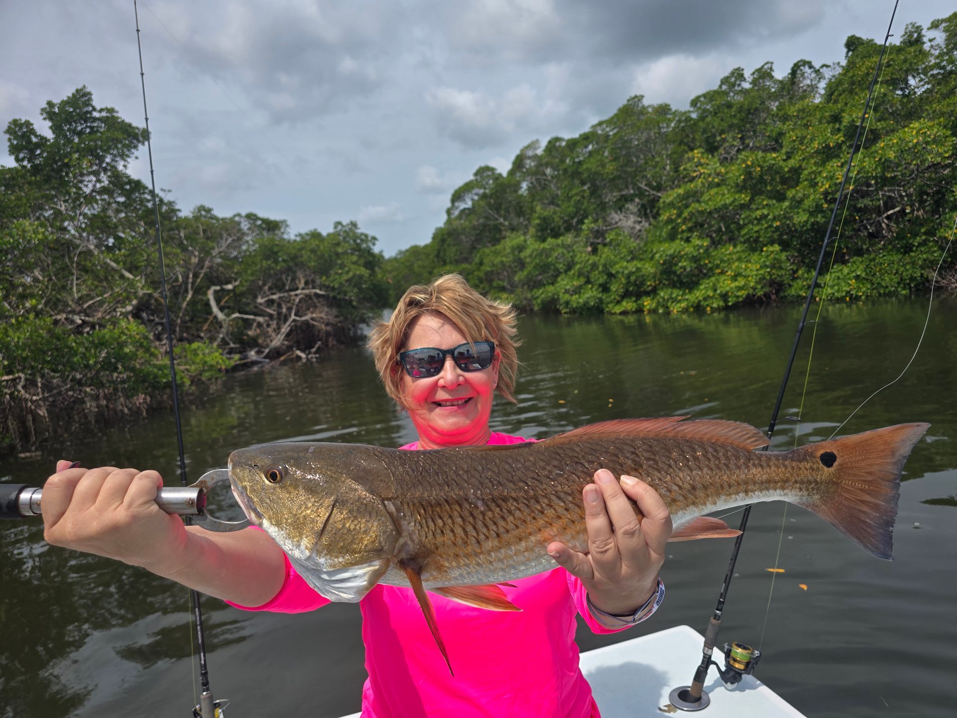Woman in pink shirt holding large redfish on a boat. Lush green mangrove backdrop.