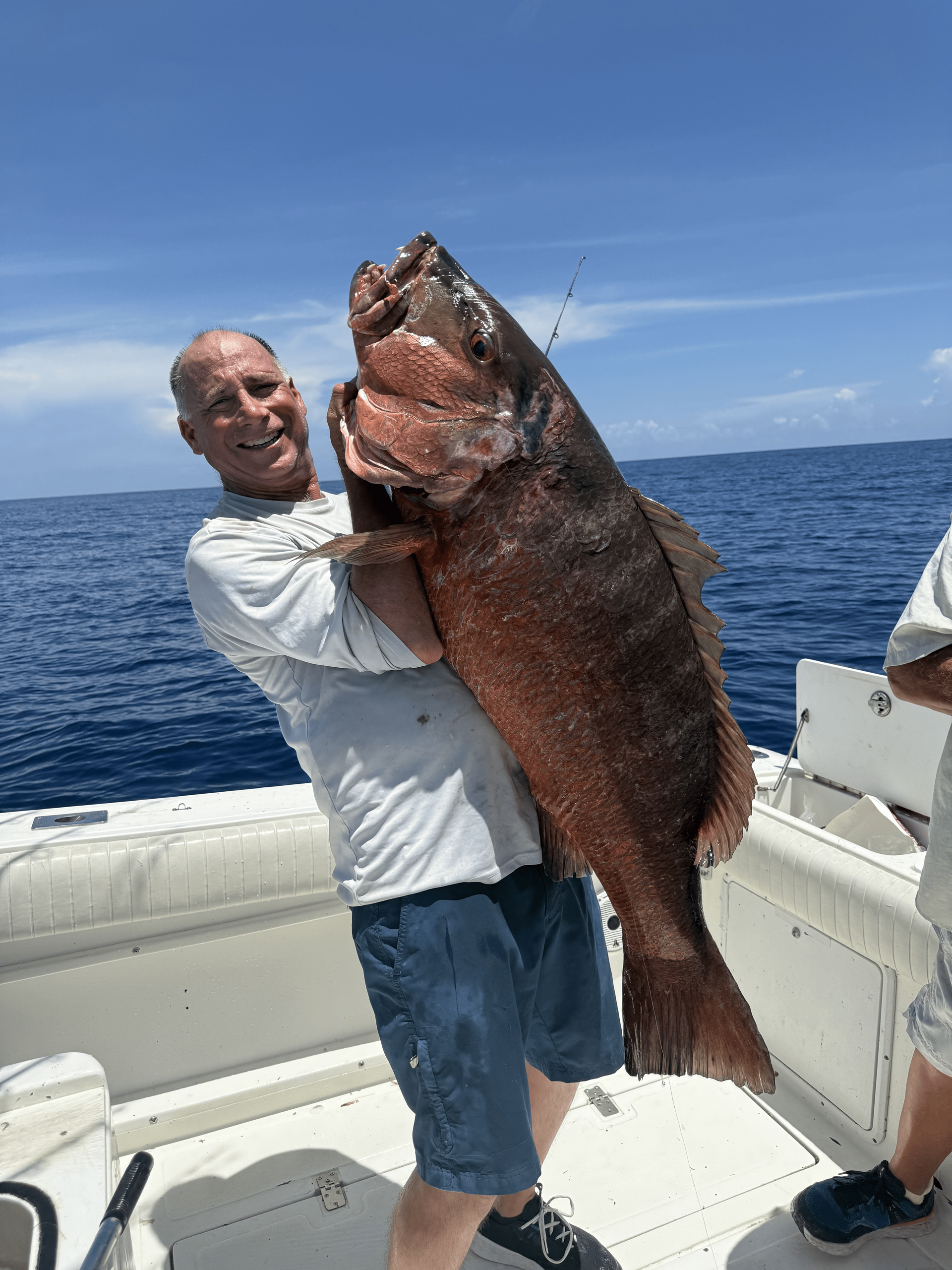 Man on boat holding huge brown fish against blue sky.