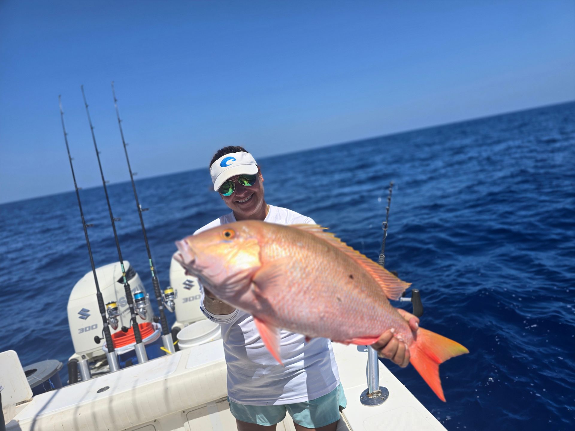Woman on boat holding a large, red snapper with orange tail, smiling at camera. Ocean in background.