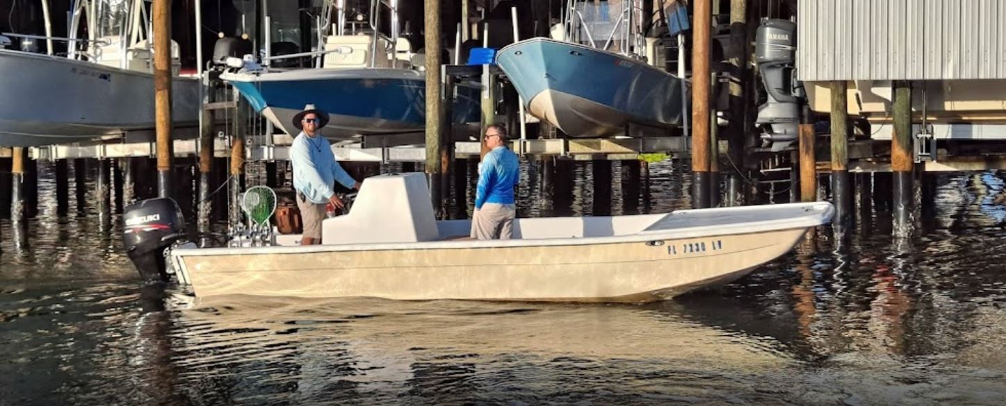 Two people on a boat in a harbor, with other boats and structures in the background.