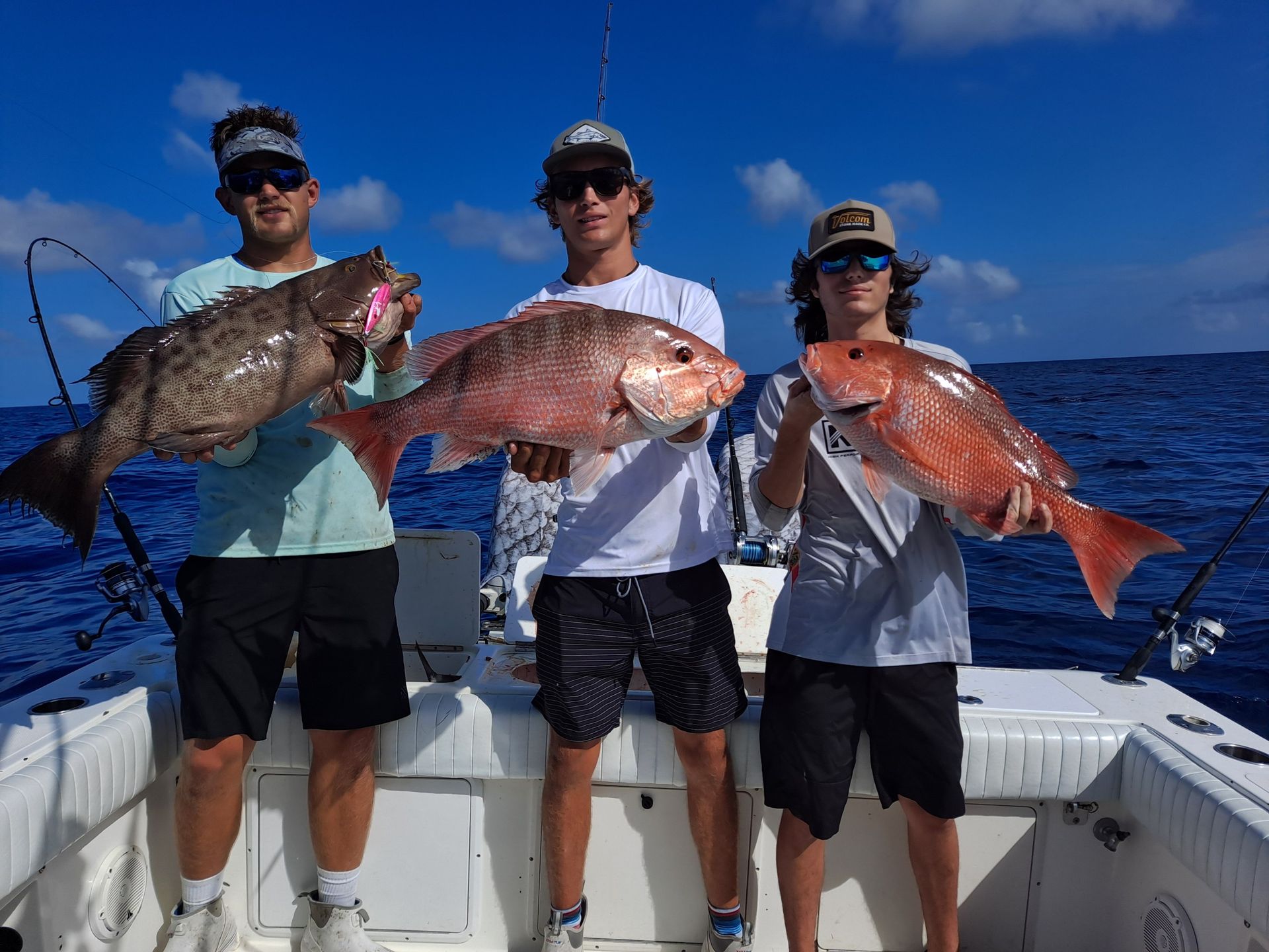 Three young men on a boat holding up large fish they caught, under a blue sky.