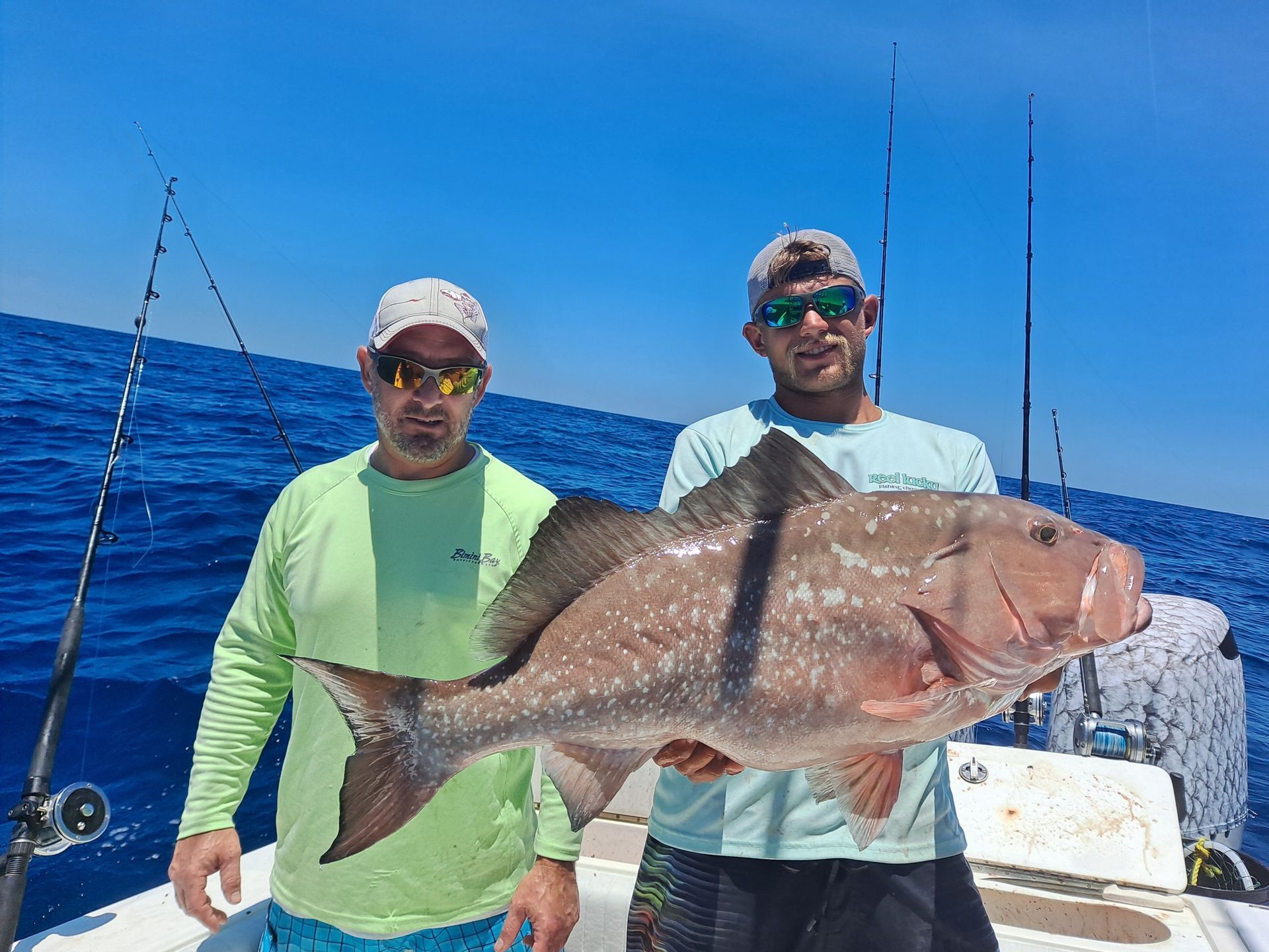 Two men on a boat holding a large, reddish-brown fish; ocean backdrop, sunny day.