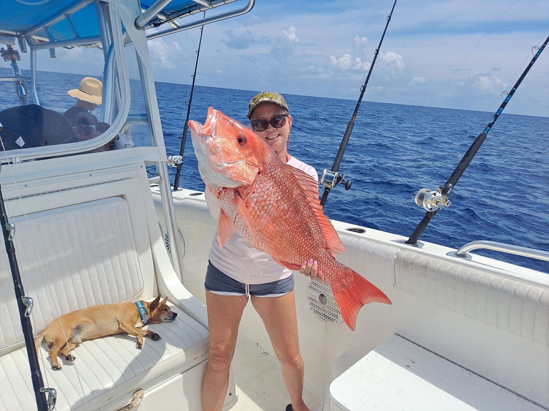 Woman on a boat holds a large, red fish; a dog lies nearby; ocean in the background.