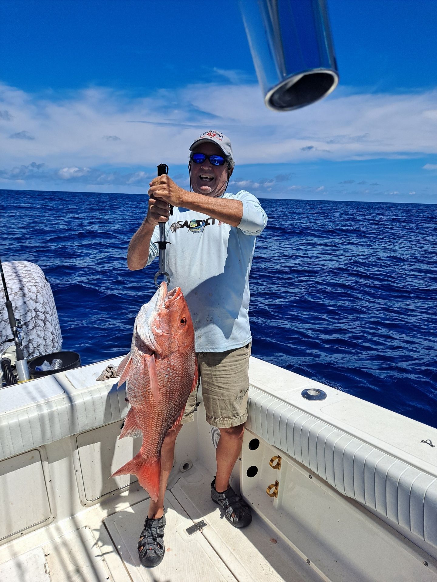 Man on a boat holds up a large red fish he caught, smiling in the sunny blue sea.