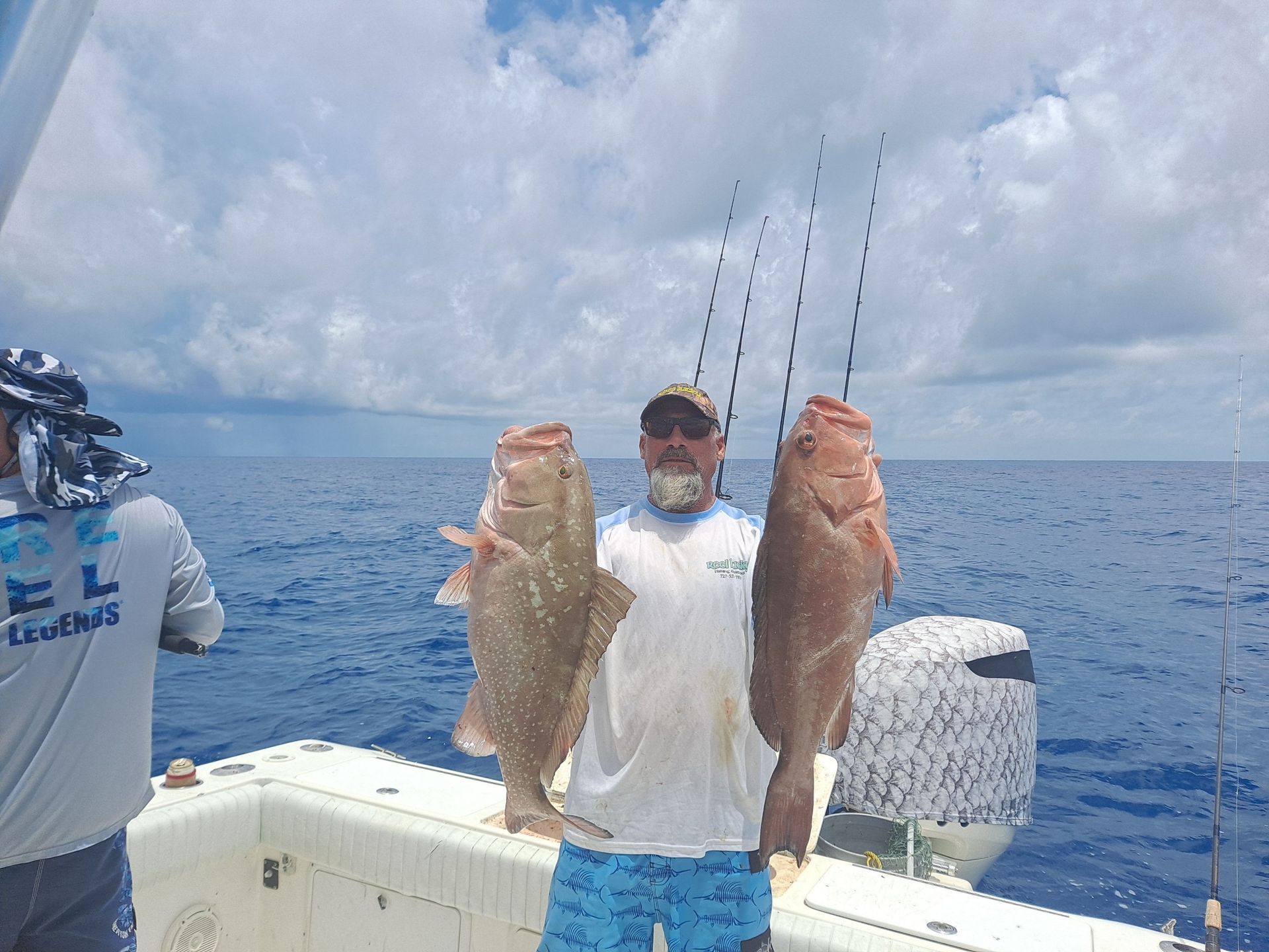 Man on boat holding two large red grouper fish. Ocean setting, blue sky.