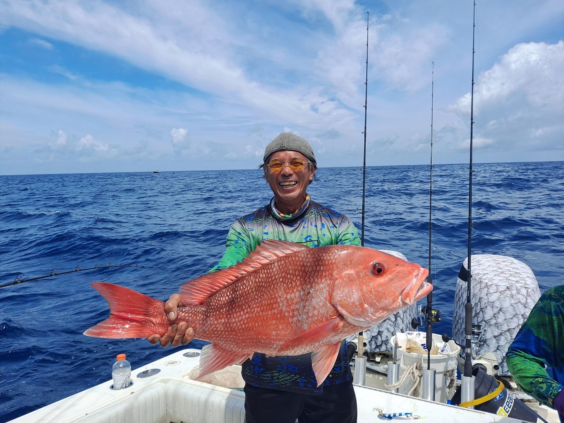 Man holding a large red snapper on a boat at sea. He's smiling, with rods visible. Blue water, cloudy sky.