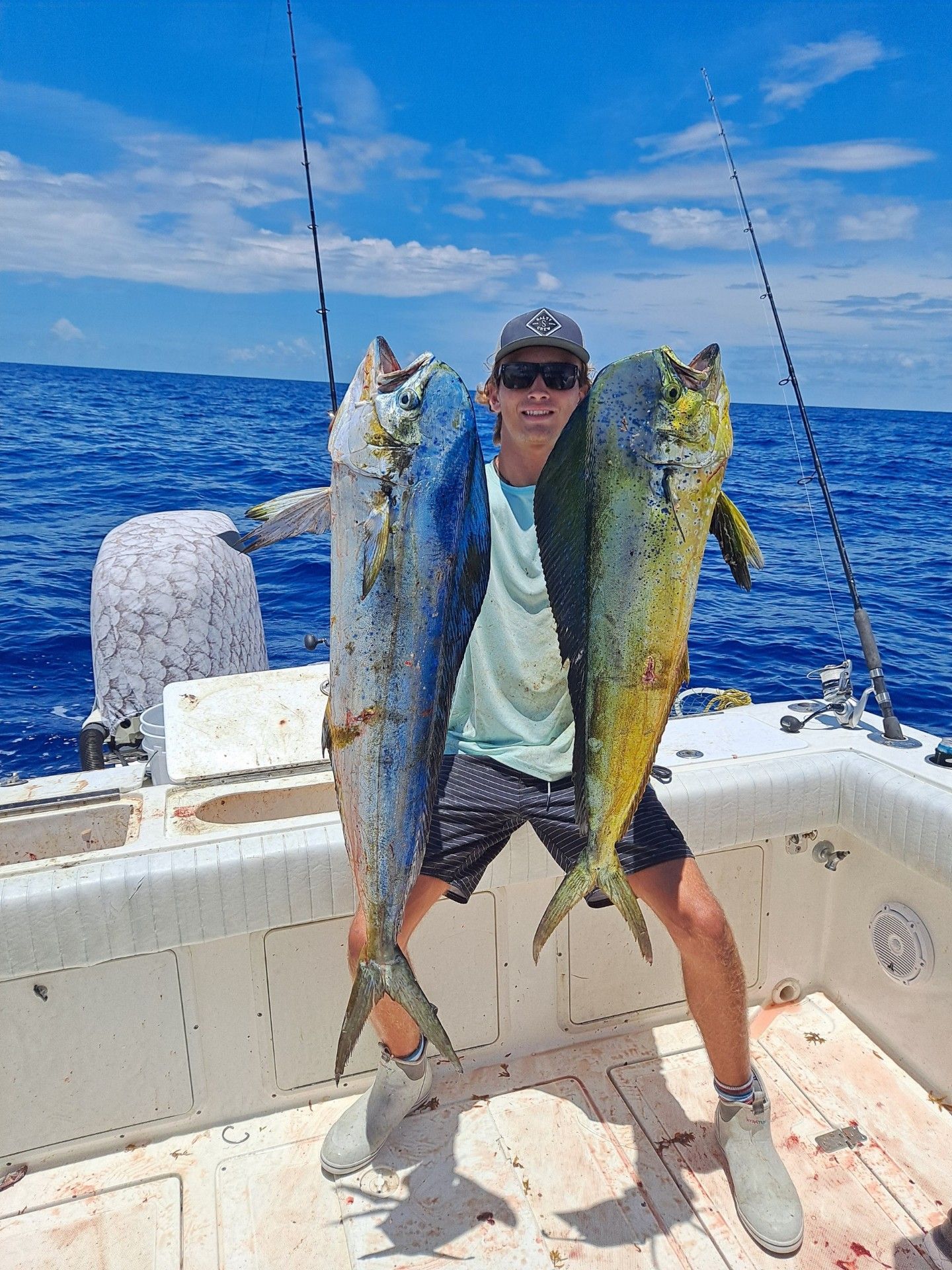 Young man on a boat, holding up two large, colorful mahi-mahi fish he caught. Ocean and blue sky background.