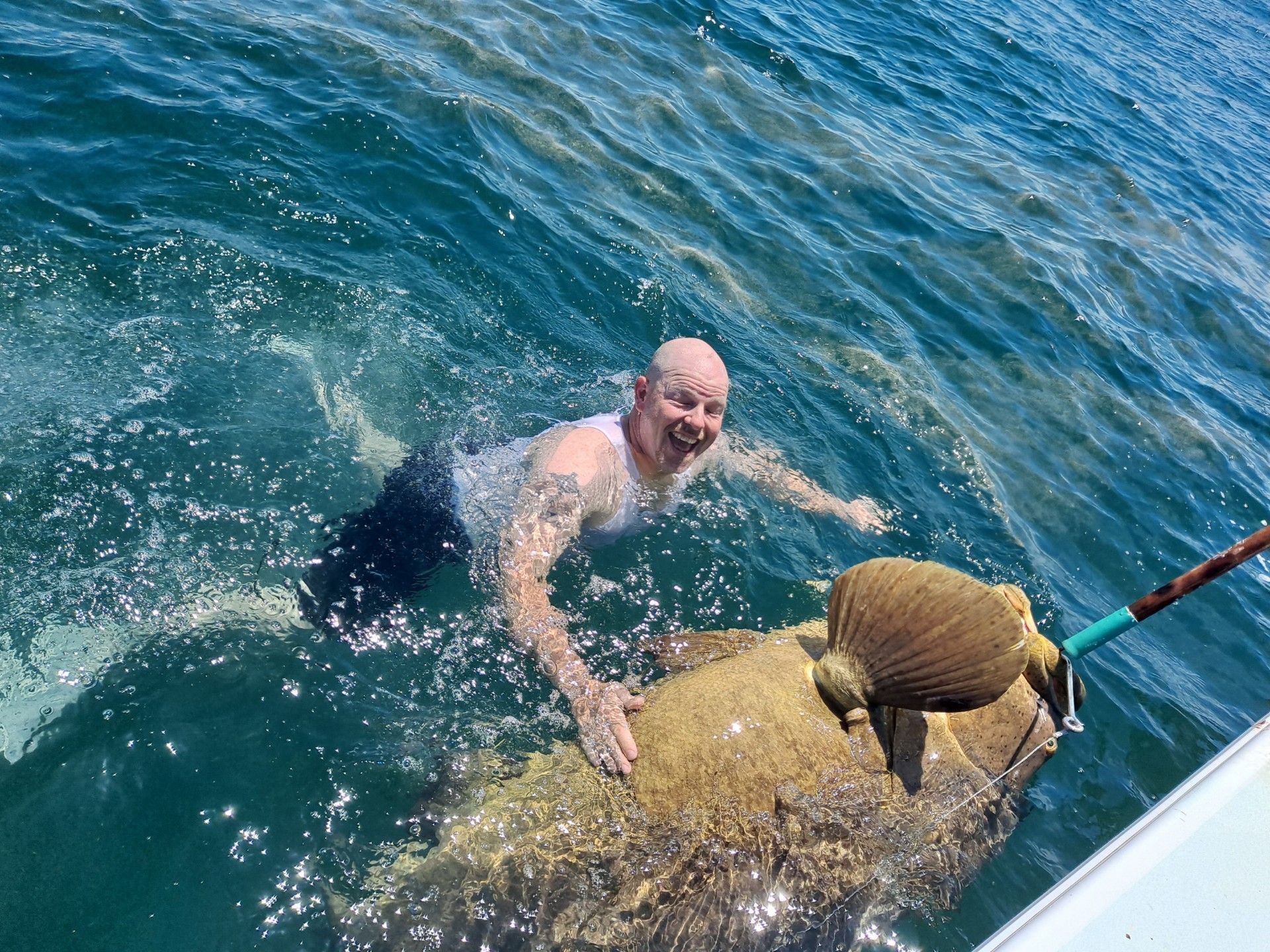 Man swimming with large fish in blue water, smiling, holding it near boat.