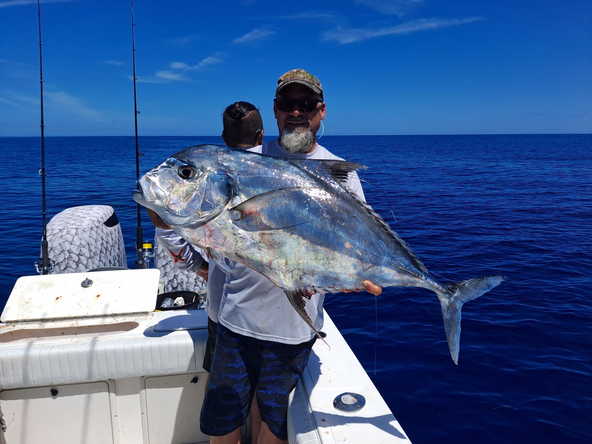 Man on boat holding a large, silver fish. Blue ocean and sky in the background.