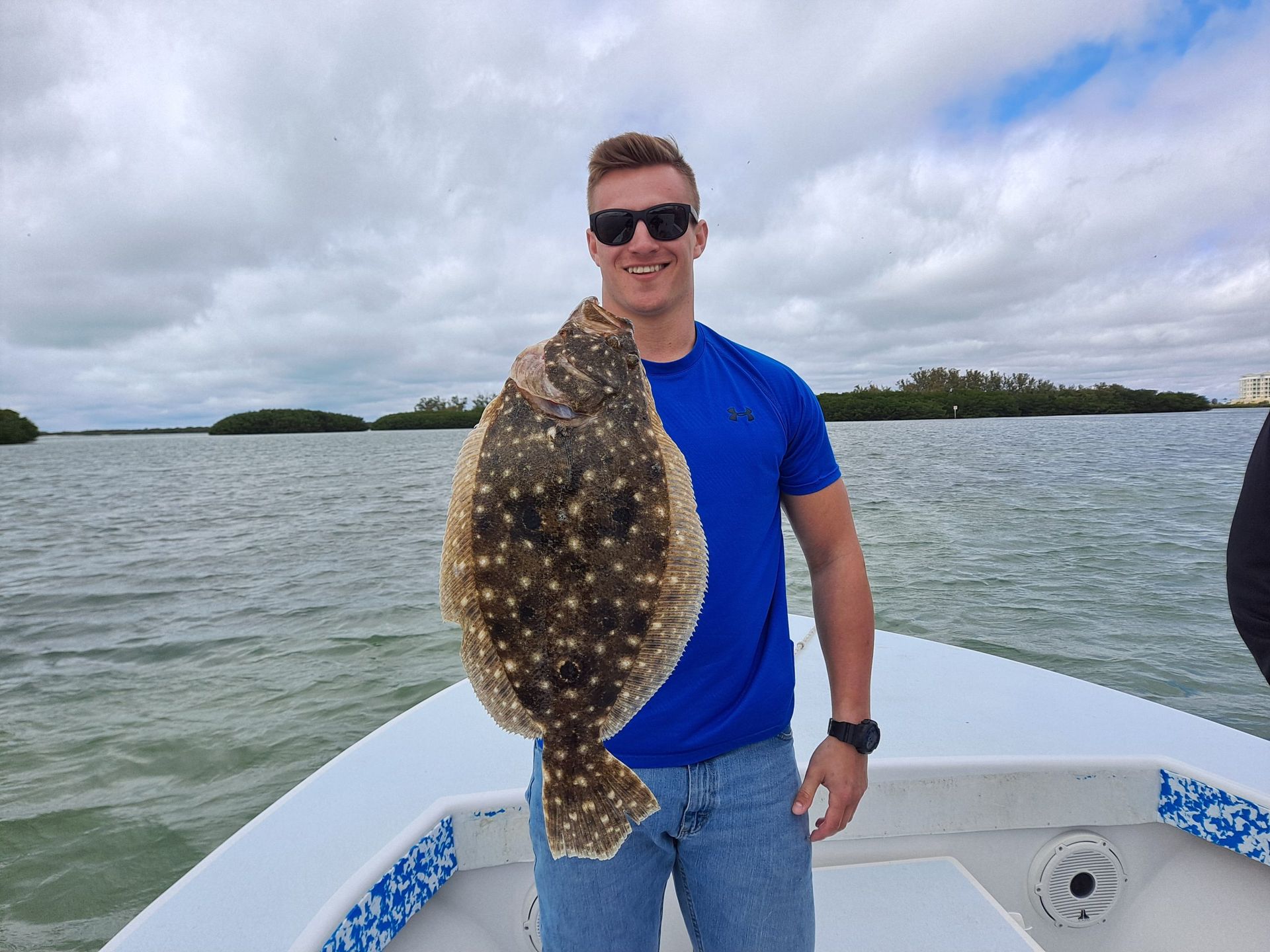 Man holding a large flounder on a boat, water and cloudy sky background.