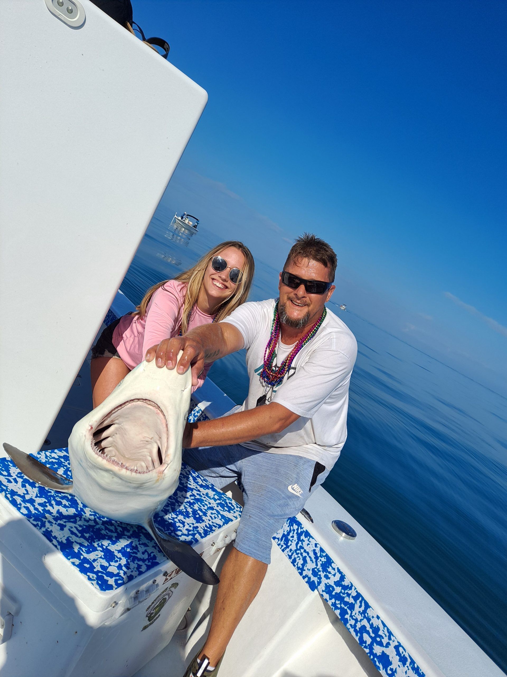 A man and woman on a boat holding a shark's mouth open. Blue sky and ocean in the background.