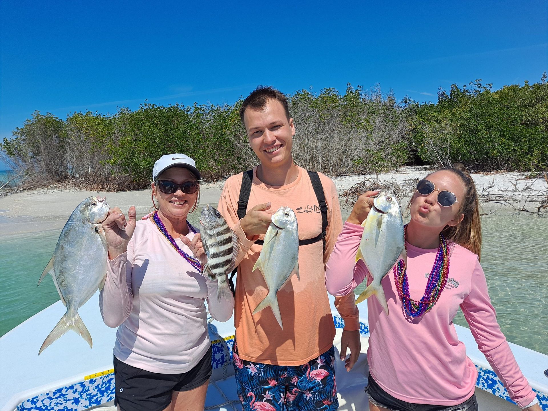 Three people smiling, holding up fish on a boat in clear water, with a blue sky and mangrove trees.