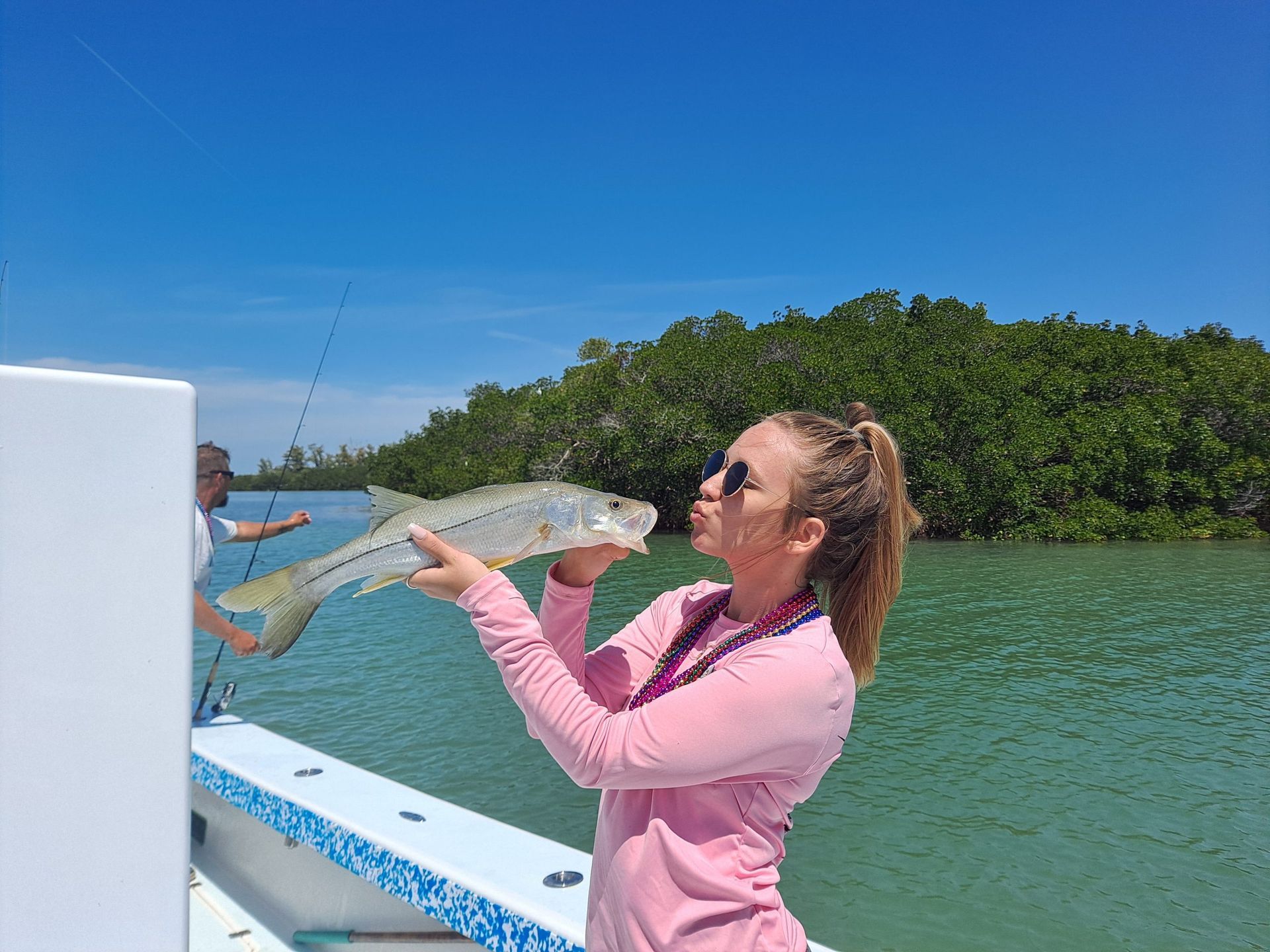 Woman on boat kisses a fish, sunny day, water, green trees in background.