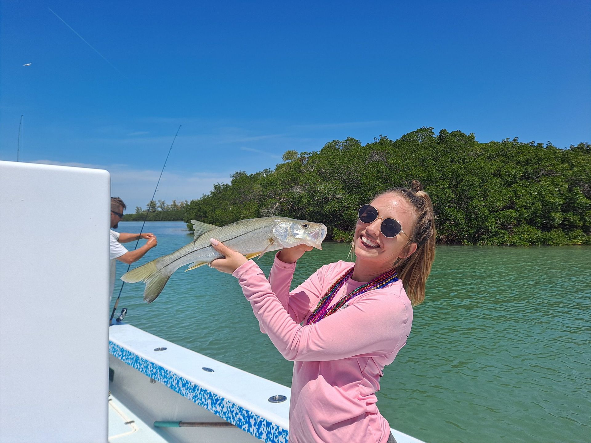 Woman on a boat smiles, holding a fish. Green water and mangroves in background, sunny day.