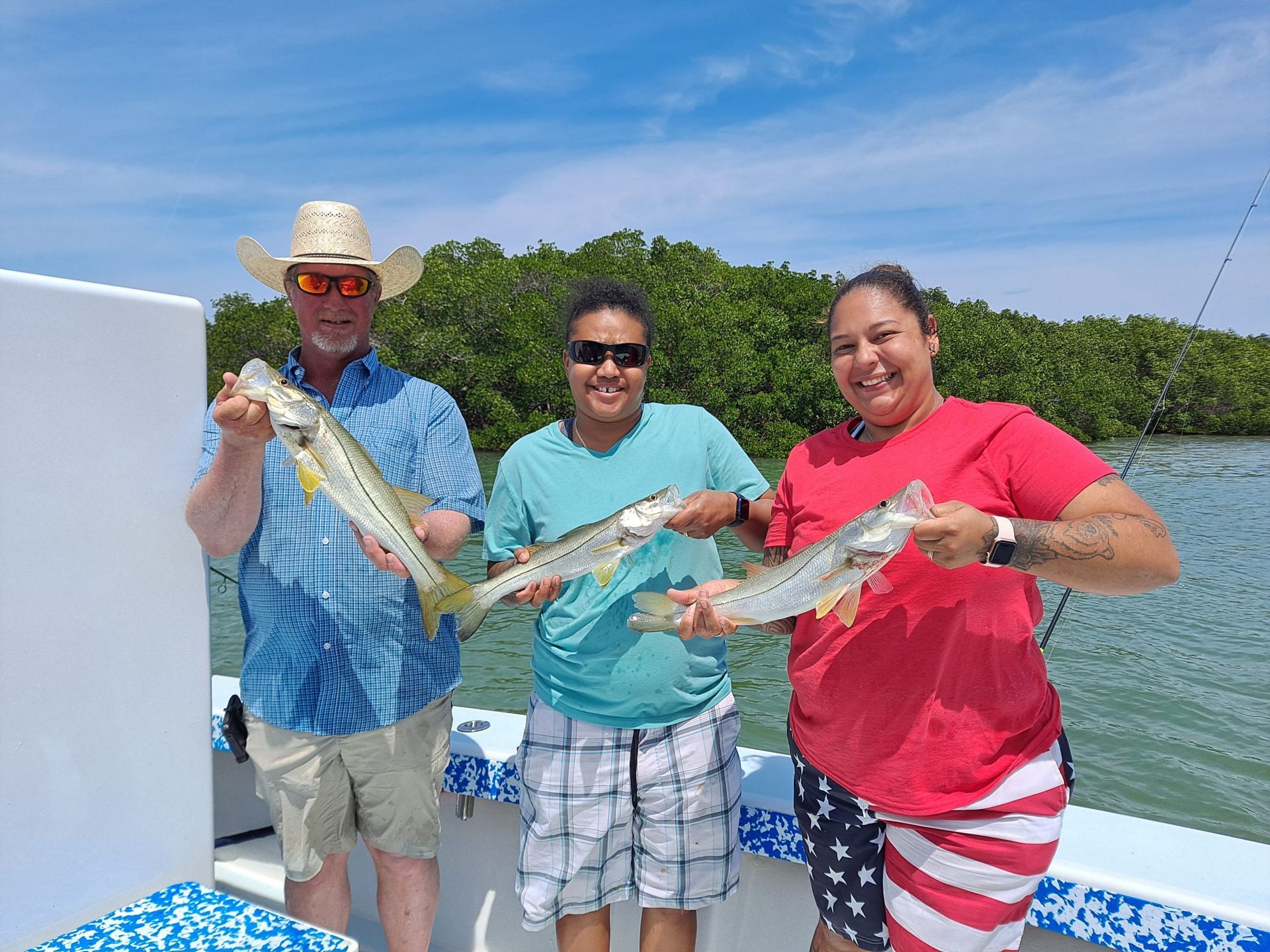 Three people smiling on a boat, holding up fish. Green water and trees in the background, blue sky.