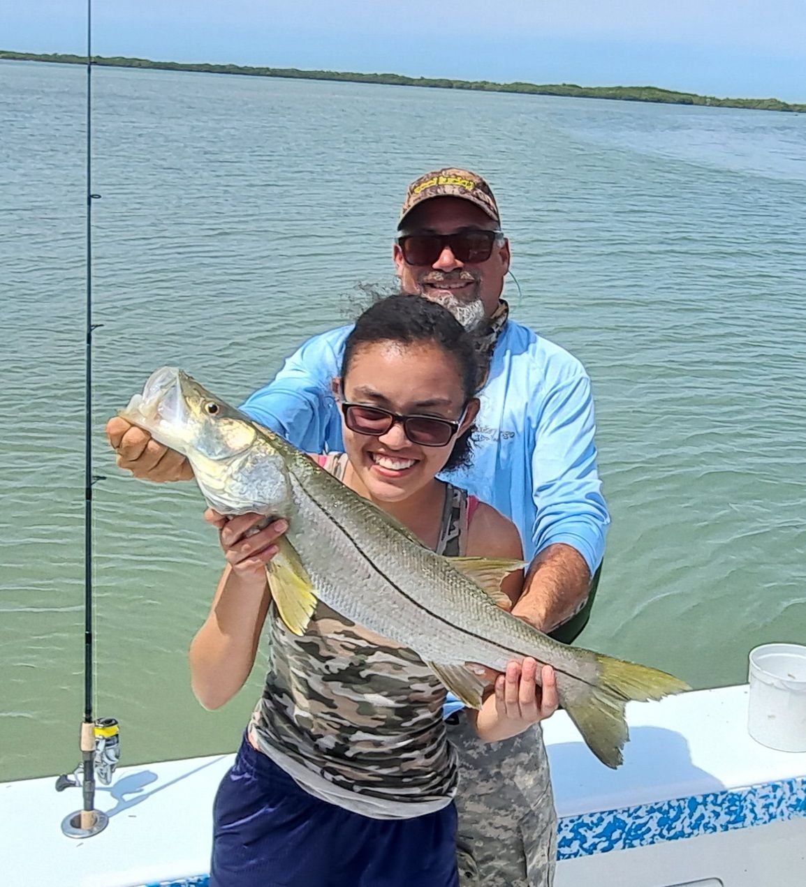 Woman and man holding a large fish on a boat, smiling. Outdoors with water and sky.