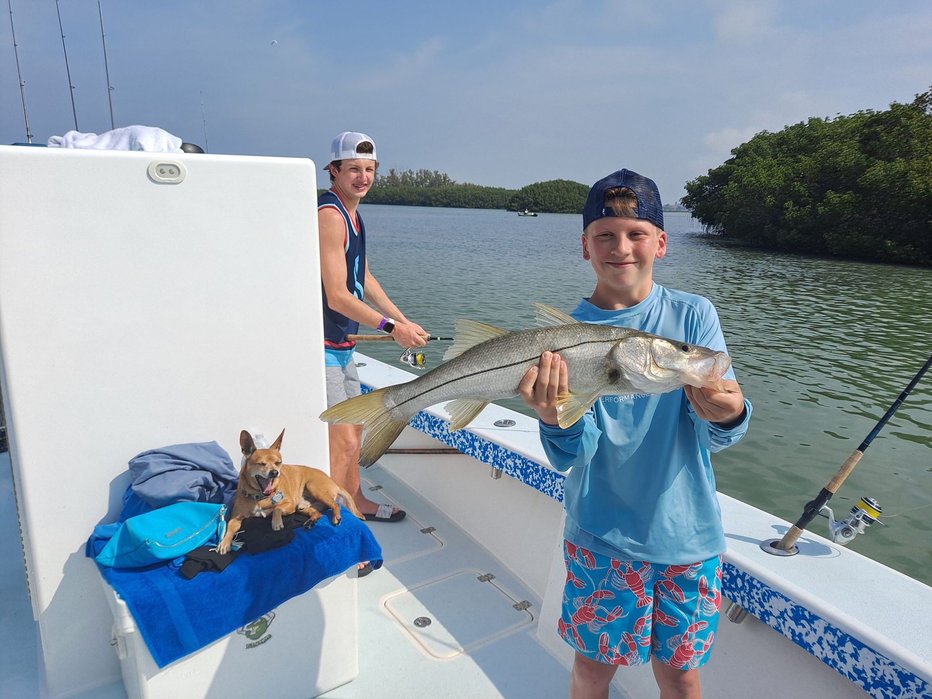 Boy on a boat holding a fish, another boy behind him fishing, a dog resting nearby, and a water background.