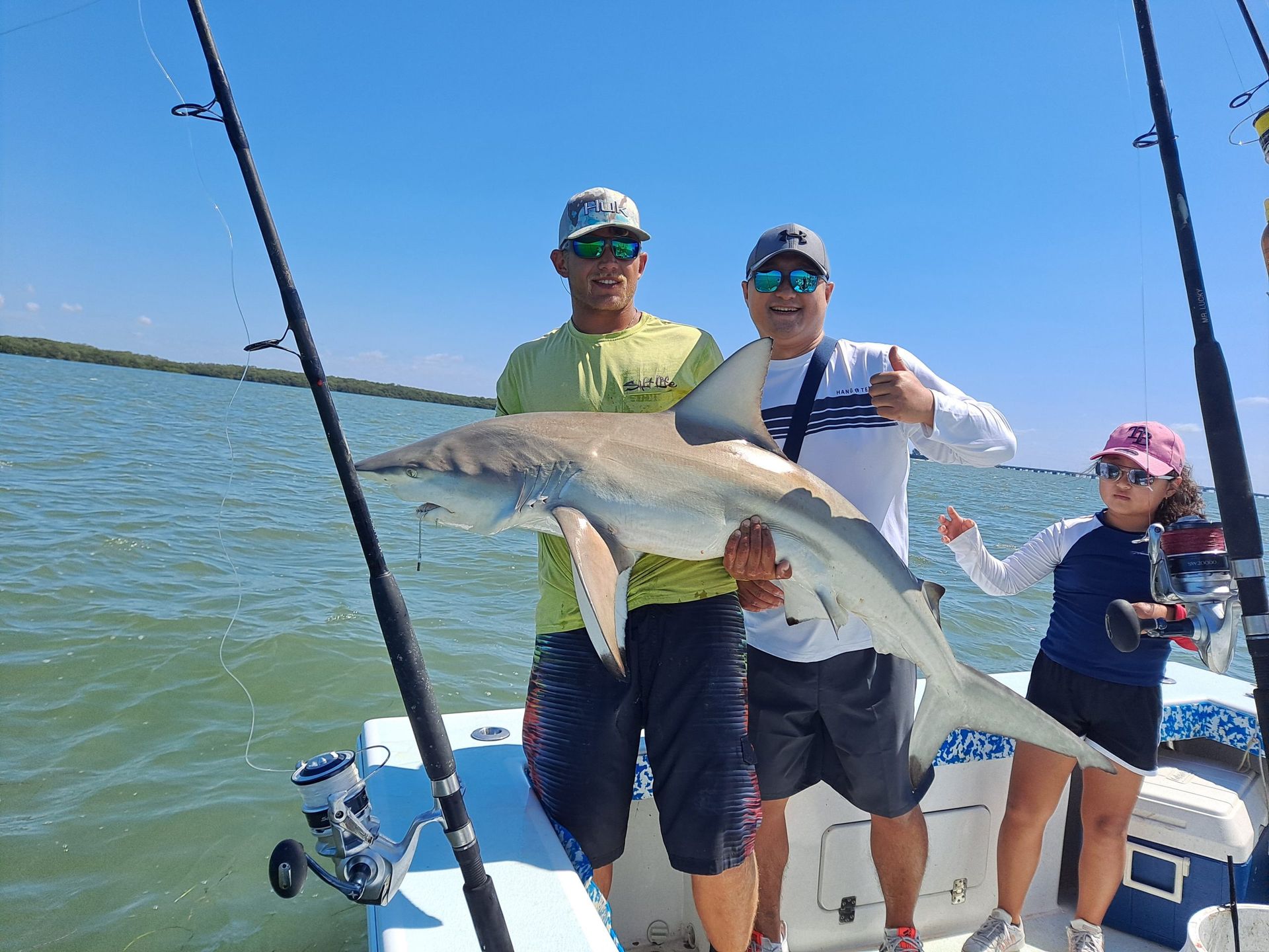 Two men and a woman on a boat holding a large shark, celebrating a successful fishing trip on a sunny day.
