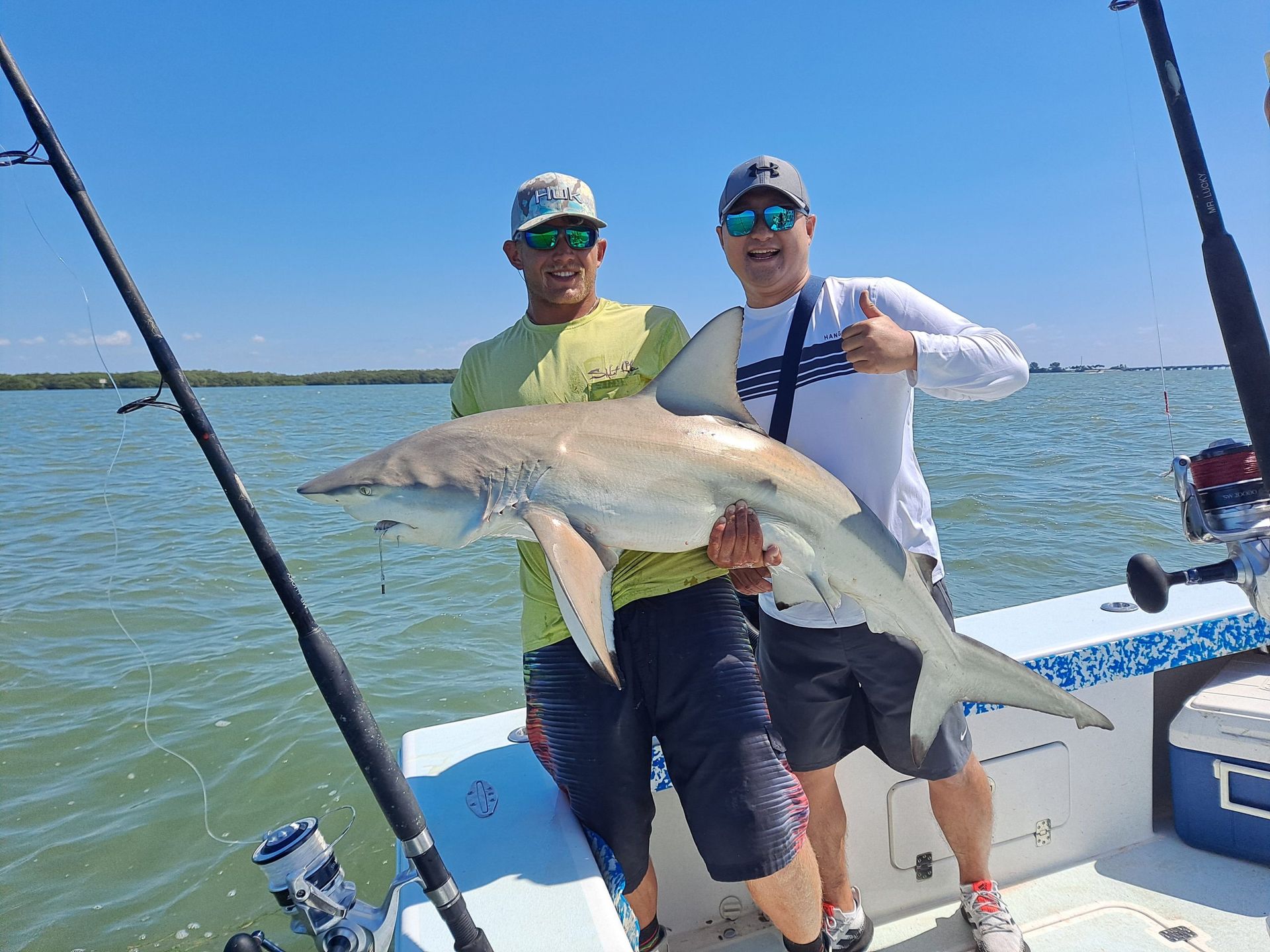 Two men on a boat hold up a large shark they caught, one gives thumbs up. Blue sky and water.