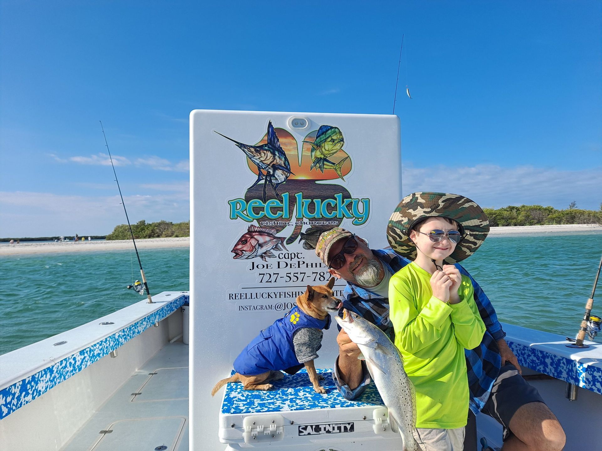 Man and boy on a boat holding a fish, dog in a blue coat. Sunny day, water, logo says 