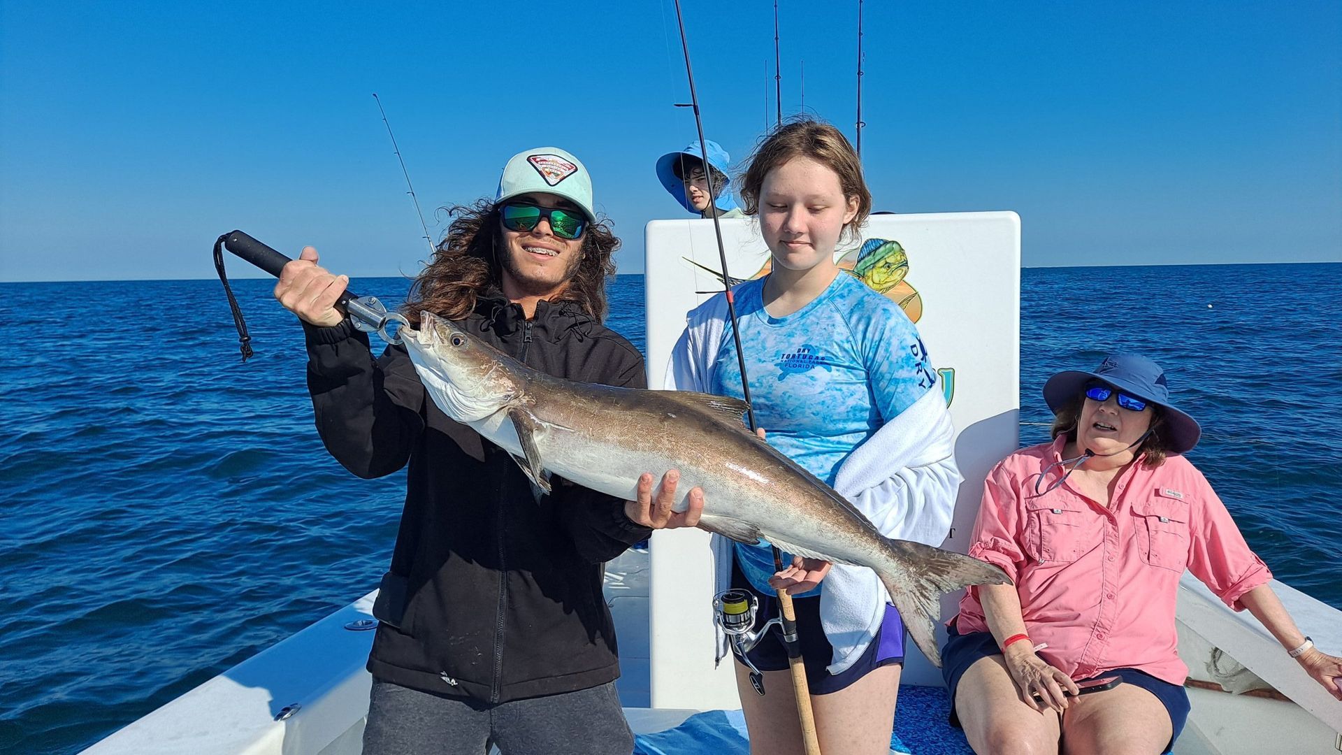 Three people on a boat with a large fish, under a blue sky. Man holding the fish, girl smiling, woman in a hat.